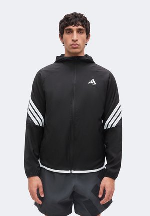 Jeune homme portant une veste noire Adidas zippée avec des bandes blanches sur les manches et un short noir, debout face à un fond uni.