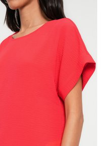 Blouse rouge texturée avec un large décolleté et des manches courtes, présentant une coupe ample qui tombe juste sous les épaules.