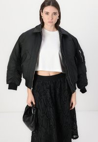 Veste aviateur noire avec fermeture éclair, poignets côtelés et épaules structurées, associée à une jupe noire texturée et un petit sac noir.