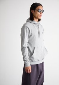 Felpa grigia con cappuccio, tasca frontale, chiusura con cordino e logo ricamato. Abbinata a pantaloni scuri e larghi.