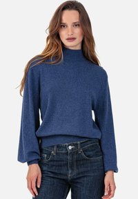 Pull en tricot bleu avec un col haut et des manches longues légèrement bouffantes, associé à un jean foncé. Le tissu semble doux et texturé.
