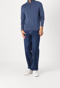 Mörkblå zip-up tröja med långa ärmar, ribbade muddar och en logotypdetalj på bröstet, tillsammans med löst sittande blå jeans och vita sneakers.