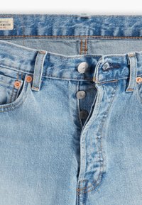 Jeans de mezclilla azul claro con cierre de botón plateado expuesto, remaches de cobre y clásicas trabillas, mostrando una textura de tela detallada y costuras.