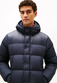 Marineblaue Daunenjacke mit Kapuze, ausgestattet mit einem gesteppten Design, Druckknöpfen und einem Logo auf der Brust. Hergestellt aus glänzendem, leichtem Material.