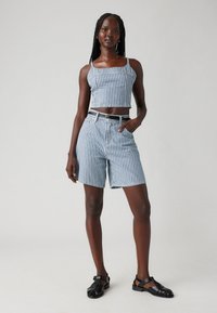 Randig blå och vit crop top med tunna axelremmar, kombinerad med matchande hög midjebyxor. Modellen bär svarta sandaler och står självsäkert.