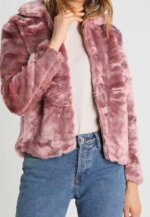 Personne portant une veste en fausse fourrure rose en peluche sur une chemise blanche et un jean bleu, avec une main touchant son cou.