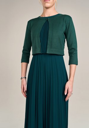Femme portant une robe plissée bleu sarcelle avec un cardigan vert court, montrant des manches 3/4 et un détail plissé au niveau de l'encolure.