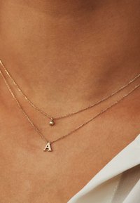 Gouden gelaagde colliers met een kleine bolpendant en een hoofdletter 'A'-bedel, weergegeven tegen een huidkleurige achtergrond.