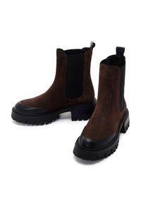 Botas Chelsea de ante marrón con paneles elásticos negros, punta redonda, suela de goma negra gruesa y tirador en la parte trasera.