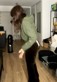 Personne portant un sweat à capuche vert et un pantalon foncé se penchant en avant dans un salon avec un sol en bois, des meubles noirs et un ventilateur tour près d'un bureau.