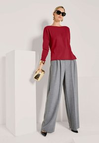 Frau in rotem Pullover, grauen weit geschnittenen Hosen, schwarzen Absätzen, großen schwarzen Sonnenbrillen, goldenen Ohrringen und Armbändern, hält eine goldene Clutch und steht neben einem weißen Sockel.