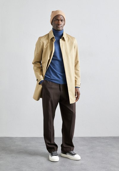 Beige trenchcoat over en blå genser med høy hals, brune stripete bukser og grå sneakers med hvite såler. Strikket lue fullfører antrekket.