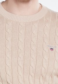 Beige Strickpullover mit Rundhalsausschnitt, der auf der linken Seite ein kleines Logo mit roten, weißen und blauen Akzenten aufweist.