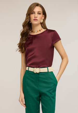 Frau mit langen, welligen Haaren, die ein kurzärmeliges Satinoberteil in Kastanienrot, grüne, hochtaillierte Hosen, einen beigen geflochtenen Gürtel und eine bunte Perlenkette trägt.