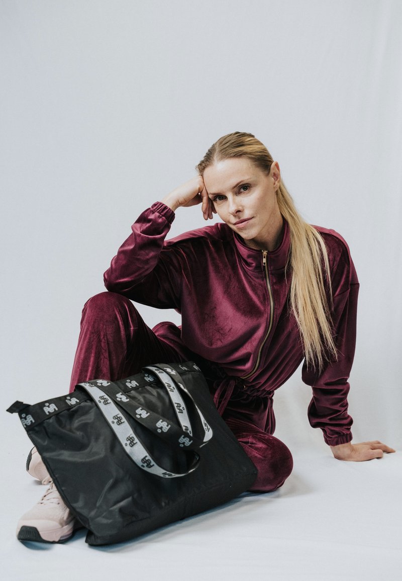 Kobieta w bordowym dresie z weluru siedząca na podłodze z czarną torbą na ramię i jasnoróżowymi sneakersami na tle jednolitego, jasnego tła.