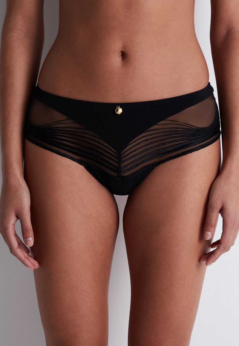 Caleçons de bikini noirs avec des panneaux en mesh transparent à rayures horizontales, une base en tissu lisse et un charme géométrique doré au centre devant.