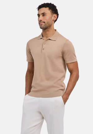 Mann steht mit Händen in den Taschen, trägt ein beigefarbenes Polohemd mit kurzen Ärmeln und weiße Hose vor einem einfarbigen Hintergrund.