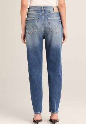 Woman wearing mid-rise, faded blue jeans with back pockets and a white sleeveless top, standing against a neutral background.