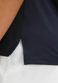 Mornarska sportska majica s teksturiranom tkaninom, blagim bočnim razdvajanjem i logotipom na rubu, preko bijele odjeće.