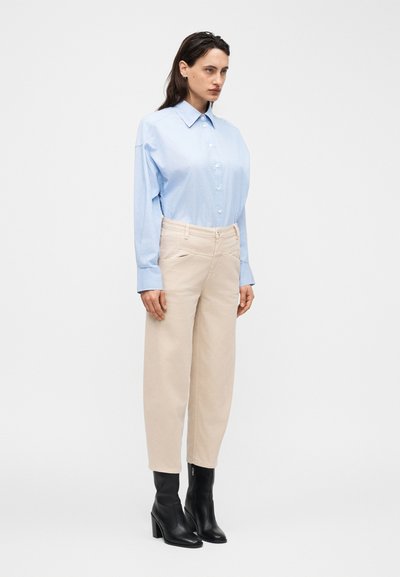 Femme portant une chemise bleu clair boutonnée, un pantalon beige à jambes larges, et des bottines noires à talons, posant devant un fond blanc uni.