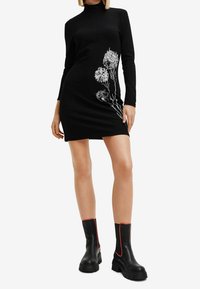 Svart långärmad klänning med en figurnära design, med vitt blommigt linjeteckning på sidan. Modellen bär svarta ankelstövlar med röda accenter.