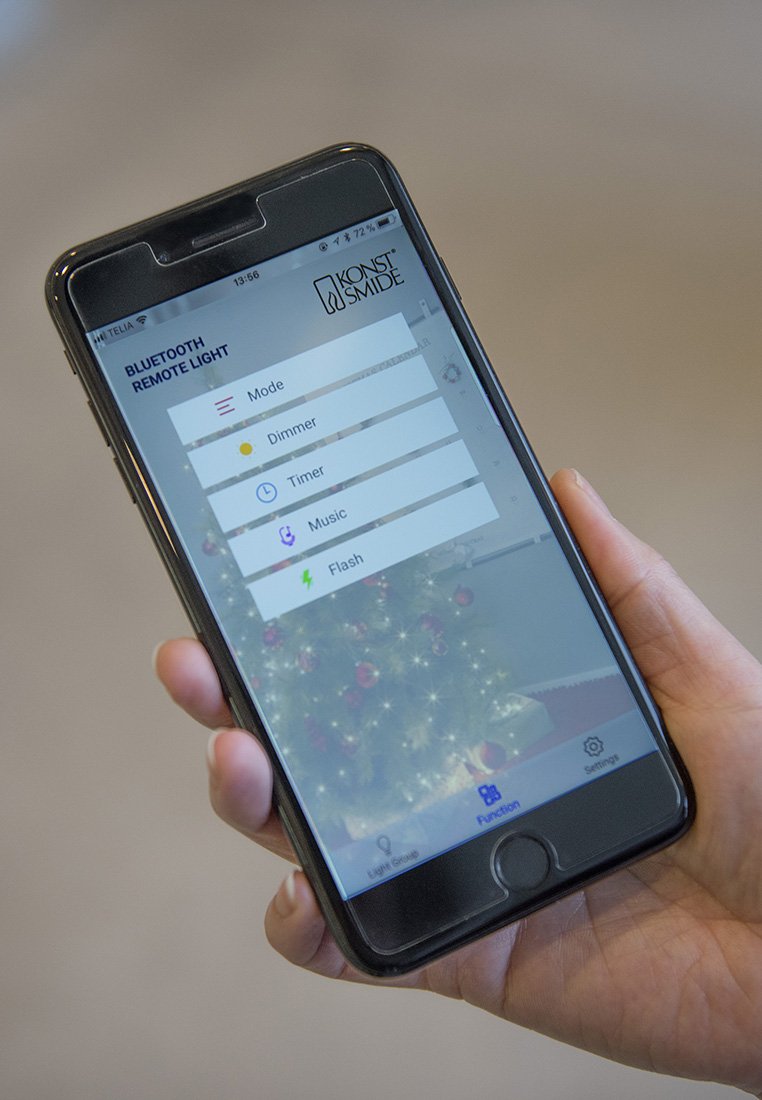 Hand die smartphone vasthoudt met een Bluetooth afstandsbediening app voor verlichting met opties voor Modus, Dimmer, Timer, Muziek en Flits, kerstboom op de achtergrond.