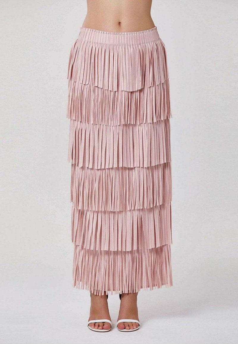 Jupe maxi frangée rose avec texture en couches, ceinture élastique et ourlet droit. Portée avec des sandales blanches à brides.