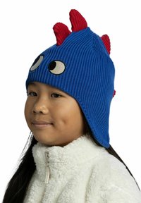 Blue knit beanie with red spikes, large white eyes, and a ribbed texture. Worn with a white fluffy jacket, featuring a high collar.
