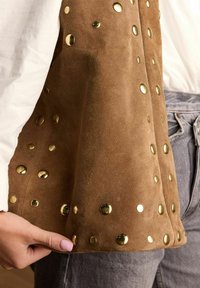 Bruine suède vest met ronde gouden studs, met een zachte textuur en een aansluitend ontwerp, gedragen over een lichtgekleurde blouse.