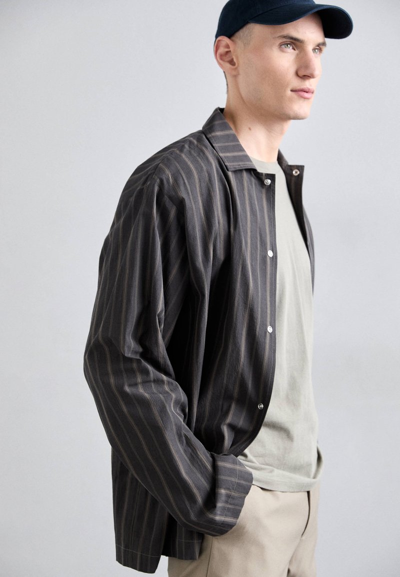 Chemise à rayures grise foncée avec des rayures claires, présentant une coupe décontractée et des manches longues, superposée à un T-shirt gris clair.