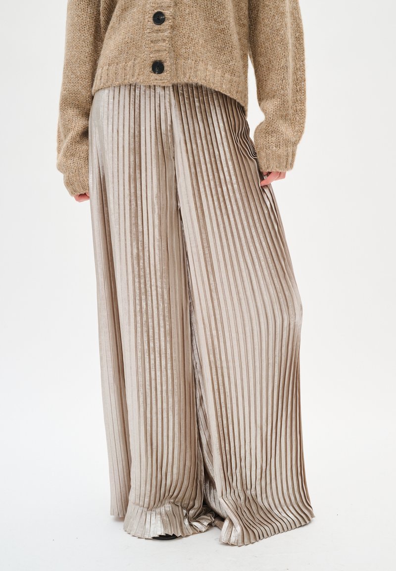 Spodnie w kant o szerokich nogawkach w jasnobeżowym kolorze, z pionowymi paskami i gładką fakturą, w zestawieniu z brązowym swetrem dzianinowym.