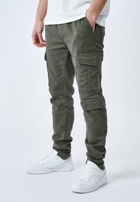 Pantaloni cargo verde oliva con multiple tasche, vestibilità affusolata e materiale misto cotone, abbinati a sneaker bianche.
