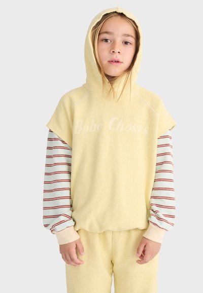 Jeune personne portant un sweat à capuche sans manches jaune pâle par-dessus un t-shirt à manches longues rayé, se tenant devant un fond clair uni.