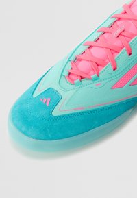 Turkis- og rosa sportssko med teksturert semsket skinn, med lisser og navnet "MESSI 10" langs siden, satt mot en hvit bakgrunn.