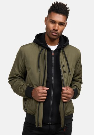 Olivgrüne Bomberjacke mit Reißverschluss, gerippten Bündchen und Taschen, über einem schwarzen Reißverschluss-Hoodie und einem weißen T-Shirt getragen. Lässige Passform.