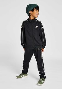 Chándal negro con franjas blancas, que incluye una chaqueta con cremallera y pantalones a juego. El niño lleva un gorro de punto verde y zapatillas deportivas verdes.