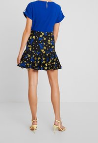 Top blu a maniche corte con dettaglio di bottoni sul retro, abbinato a una gonna nera a campana con stampe floreali gialle e accenti di foglie blu. Tacchi gialli.