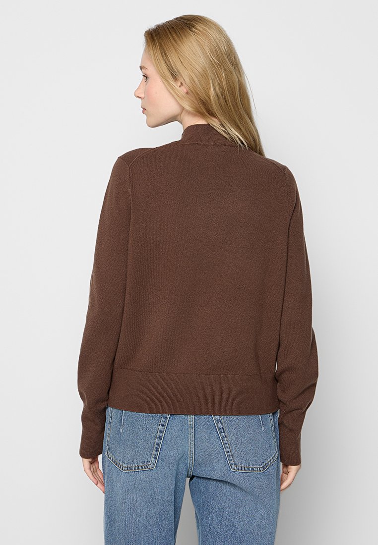 Braun gestrickter Pullover mit hohem Kragen, gerippten Bündchen und Saum; kombiniert mit hellblauen hoch taillierten Jeans; glatte Textur.