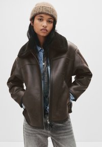 Mladá žena nosící chlupatou béžovou čepici, tmavě hnědou koženou bundu s kožešinou, modrou pruhovanou košili a šedé džíny, stojící s rukama v kapsách.