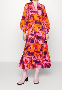 Lange, gelaagde jurk in levendige roze, oranje en paarse patronen. Heeft opgezette mouwen en een V-halslijn, en wordt gedragen met kniehoogte roze laarzen.