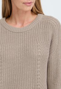 Beige gestrickter Pullover mit rundem Ausschnitt, der ein strukturiertes Muster und gerippte Kanten aufweist, und einen weichen sowie gemütlichen Look vermittelt.