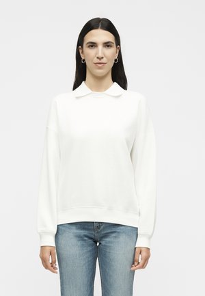Femme aux longs cheveux foncés portant des boucles d'oreilles anneaux argentés, un sweat-shirt blanc à col et un jean bleu, debout devant un fond clair uni.