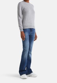 Osoba oblečená v světle šedém svetru s dlouhým rukávem, modrých džínech bootcut a bílých chunky teniskách, stojící na bílém pozadí.