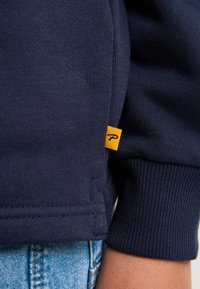 Felpa di un blu scuro con una texture a coste, polsini elastici e un piccolo etichetta gialla con un logo sul lato. Indossata con jeans azzurri chiari.