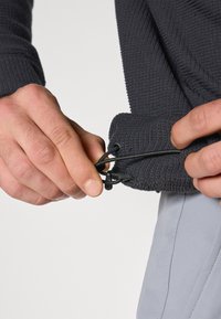Suéter texturizado gris oscuro con cordón en el dobladillo, sostenido por una mano. Una prenda gris claro es parcialmente visible debajo.