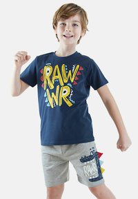 Marineblå t-shirt med gul "RAW" og "WR" tekst, røde og blå detaljer; lysegrå shorts med et dinosaur-motiv og farverige pigge.