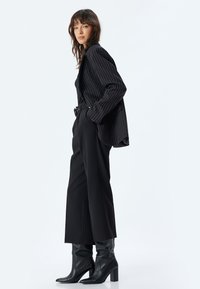 Blazer noir à rayures verticales porté sur un haut noir, associé à un pantalon noir ample et des bottes noires jusqu'aux genoux. Textures lisses et coupe structurée.
