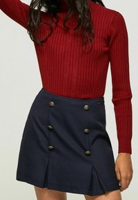 Red ribbed turtleneck sweater paired with a navy mini skirt featuring dual side pleats and six decorative gold-tone buttons.