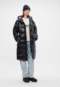 Melna pūkaina jaka ar spīdīgu apdari, ar rāvējslēdzēju un kapuci. Valkāta virs gaišas krāsas džempera un brīvā stila džinsiem, kopā ar lielām zābaku kurpēm.