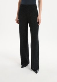 Pantaloni neri scintillanti con design svasato, caratterizzati da una vita alta e una texture morbida. Indossati con scarpe nere a punta.